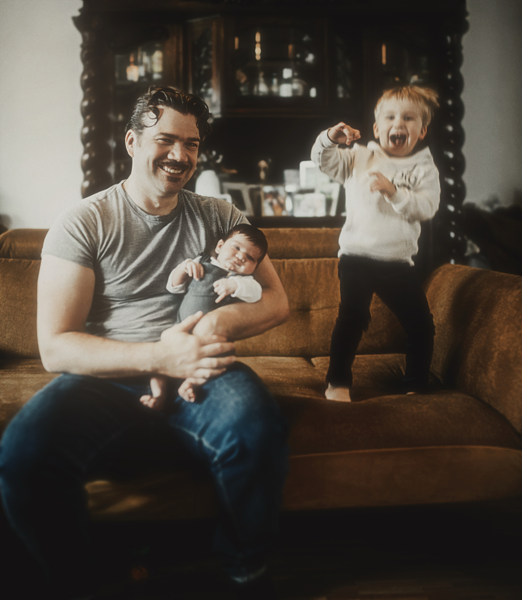 Vater mit Baby auf dem Arm, älterer Bruder springt lachend auf dem Sofa
